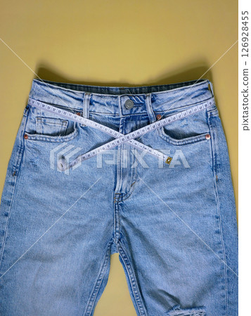 Female blue jeans with measuring tape flat lay on a yellow background. Losing weight, overweight and healthy lifestyle concept Female blue jeans with measuring tape flat lay on a yellow background. Losing weight, overweight and healthy lifestyle concept 126928455