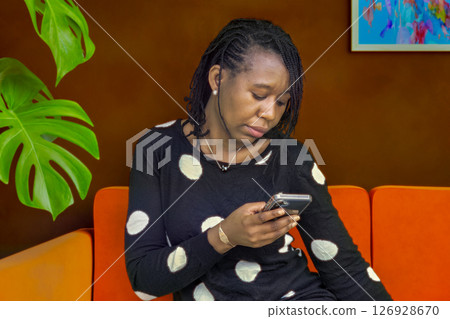 African Woman Texting on Smartphone While Sitting on Couch Indoors African Woman Texting on Smartphone While Sitting on Couch Indoors 126928670