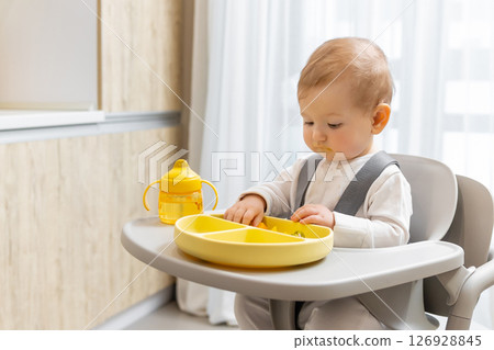 Sweet little baby boy sitting on his chair and eating something from the plate 126928845