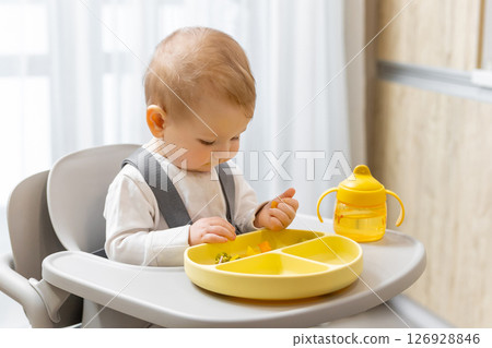 Sweet little baby boy sitting on his chair and eating something from the plate Sweet little baby boy sitting on his chair and eating something from the plate 126928846