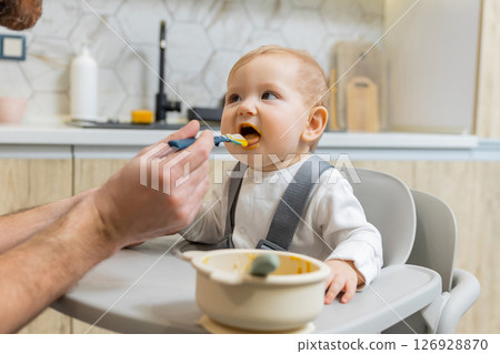 Dad feeding his little baby boy while kid sitting in his chair 126928870