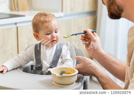 Dad feeding his little baby boy while kid sitting in his chair 126928874