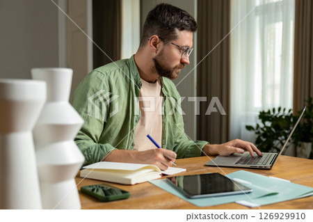 Bearded man reviewing notes on desk while working online with laptop and papers 126929910