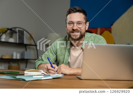 Bearded man reviewing notes on desk while working online with laptop and papers Bearded man reviewing notes on desk while working online with laptop and papers 126929920