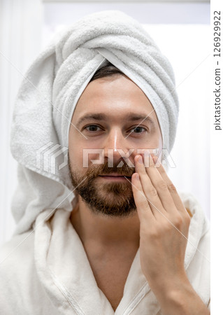 Bearded man in towel on head applying cream on his face Bearded man in towel on head applying cream on his face 126929922