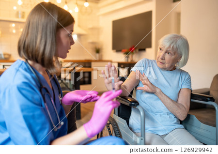 Nurse in uniform preparing medicine for an injection, elderly patient looking scared Nurse in uniform preparing medicine for an injection, elderly patient looking scared 126929942
