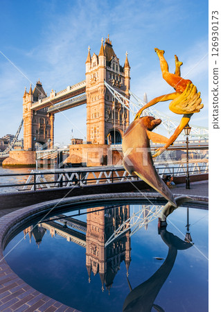 Girl with a Dolphin - a vibrant statue features a girl joyfully interacting with a dolphin, set against the iconic Tower Bridge in London. The sun illuminates the scene, creating a picturesque moment. 126930173