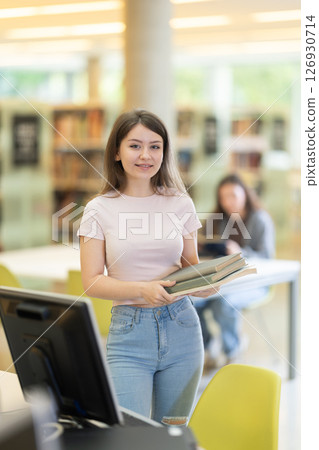 Young female student stands in library with stack of books 126930714