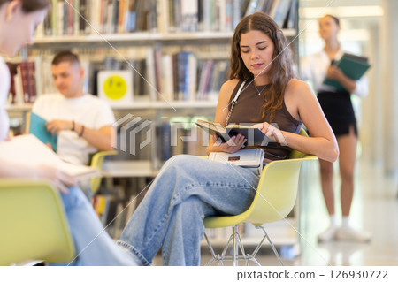 Female student sitting with book in library on shelving background Female student sitting with book in library on shelving background 126930722