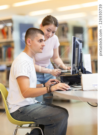 Male student works on computer in library and talks with female friend. Male student works on computer in library and talks with female friend. 126930739