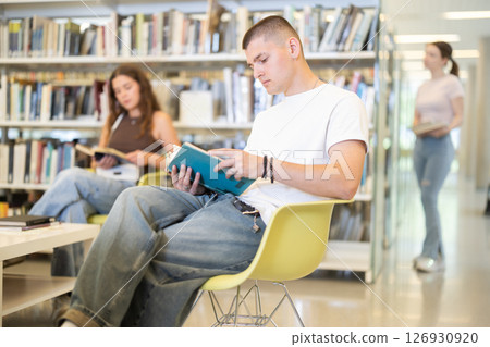 Male student sitting with book on chair in library 126930920