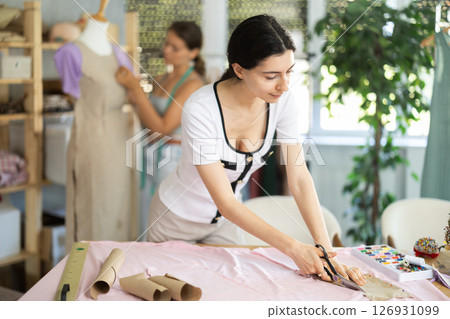 Female tailor cutting fabric while teammate working with mannequin Female tailor cutting fabric while teammate working with mannequin 126931099
