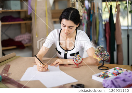 Female tailor drawing sketches of custom garment by hand in studio Female tailor drawing sketches of custom garment by hand in studio 126931130
