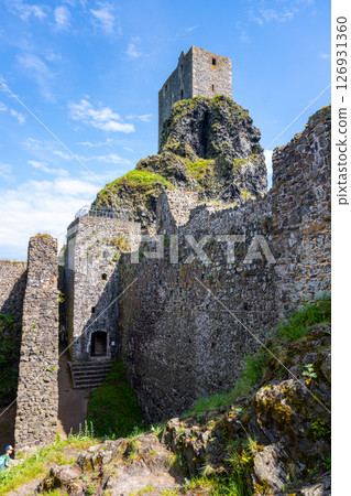 Visitors wander among the majestic ruins of Trosky Castle, perched on a rocky hill, surrounded by lush greenery and blue skies in Bohemian Paradise, Czechia, during a sunny afternoon. 126931360