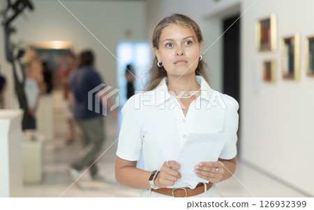 Woman with sheet piece of paper in hands view pictures and icons in museum hall Woman with sheet piece of paper in hands view pictures and icons in museum hall 126932399
