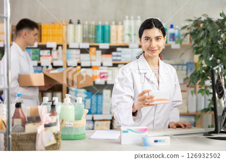 Female pharmacist in uniform looking for medicine in pharmacy using computer. In background, male pharmacist places Female pharmacist in uniform looking for medicine in pharmacy using computer. In background, male pharmacist places 126932502