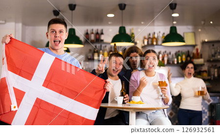 Company of young adult enthusiastic fans waving flag of Denmark and supporting national team with beer in pub 126932561