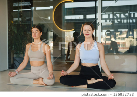 Two beautiful girls doing yoga in lotus pose. Two girls meditating at yoga sitting in lotus pose in the fitness center Two beautiful girls doing yoga in lotus pose. Two girls meditating at yoga sitting in lotus pose in the fitness center 126932758