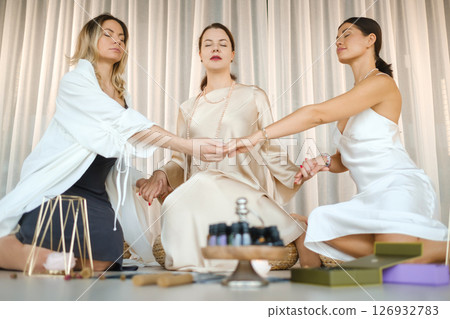 Three girls in light-colored dresses sit on the floor and hold hands in a practical session on health, beauty and creativity. Three beautiful girls holding hands Three girls in light-colored dresses sit on the floor and hold hands in a practical session on health, beauty and creativity. Three beautiful girls holding hands 126932783