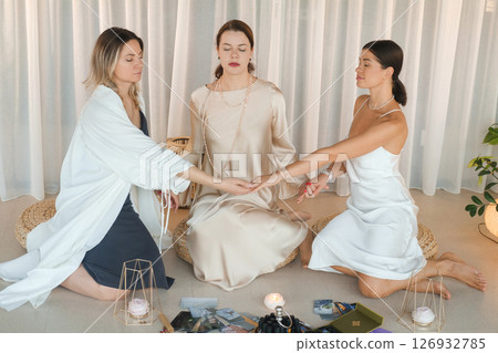 Three girls in light-colored dresses sit on the floor and hold hands in a practical session on health, beauty and creativity. Three beautiful girls holding hands Three girls in light-colored dresses sit on the floor and hold hands in a practical session on health, beauty and creativity. Three beautiful girls holding hands 126932785