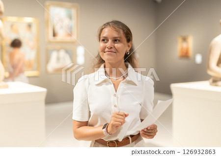 Girl visitor looks with interest at the sculpture in the museum hall, holding an information booklet in her hands 126932863