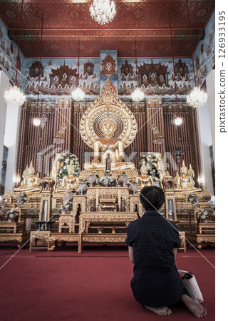 A woman is sitting in front of a golden Buddha statue in a temple. A woman is sitting in front of a golden Buddha statue in a temple. 126933195