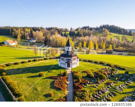 An aerial perspective showcases the white octagonal Tesarov Chapel nestled in the lush greenery of Korenov, surrounded by colorful trees and a peaceful cemetery, creating a serene landscape. 126935166