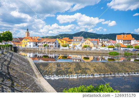 The tranquil Otava River flows through Susice, reflecting colorful buildings under a bright blue sky filled with fluffy clouds. This scenic spot captures the charm of a quaint Czech town. 126935170