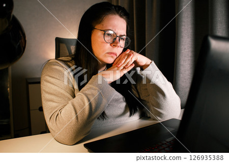 Young woman with eyeglasses working on laptop at home late at night, using modern technologies and getting online education. Remote learning concept. Online university. Focus on process. Young woman with eyeglasses working on laptop at home late at night, using modern technologies and getting online education. Remote learning concept. Online university. Focus on process. 126935388