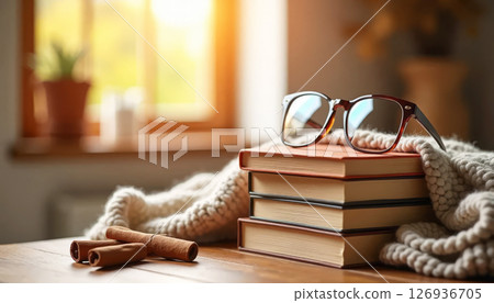Stack of books, glasses, cinnamon, and blanket on wooden table in warm sunlight. Autumn reading and cozy home atmosphere. 126936705