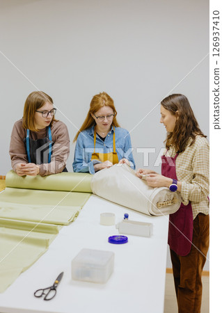 Three female seamstresses fashion designers stand at a table with roll of fabric and negotiate. 126937410