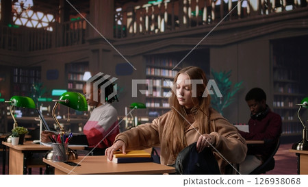 Female college student reads and writes detailed notes for a new course, studying political science for upcoming lecture in a library setting at university. Girl consults database. Camera B. Female college student reads and writes detailed notes for a new course, studying political science for upcoming lecture in a library setting at university. Girl consults database. Camera B. 126938068