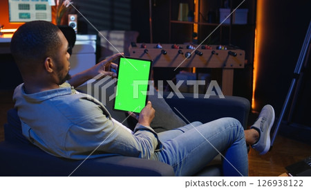 Black remote worker looking at green screen on his device at home, relaxing in the living room and solving freelancing tasks. Guy being casual in trendy apartment, teleworking. Camera A. 126938122