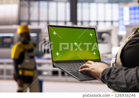 African american factory worker holds laptop with green screen display, overseeing automated production line. The industrial plant integrates robotics, conveyor belts and smart manufacturing systems. 126938246