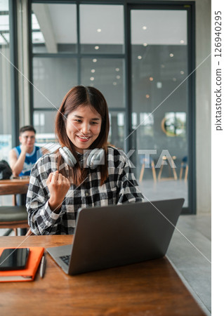 Smiling woman in plaid shirt celebrates success on laptop in a modern cafe, headphones around neck, expressing joy. 126940295