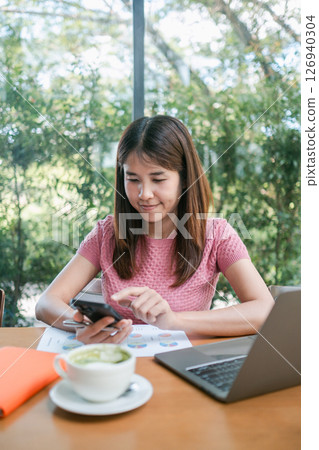 A young woman in a pink shirt uses her smartphone while working on a laptop in a bright cafe environment. A young woman in a pink shirt uses her smartphone while working on a laptop in a bright cafe environment. 126940304