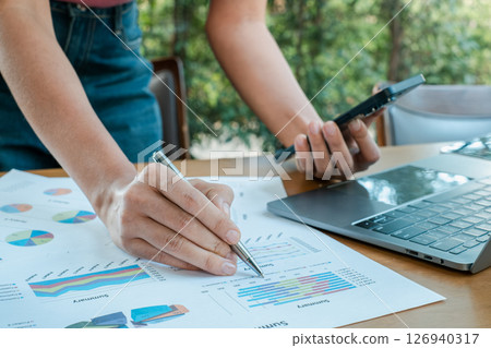 Close-up of hands reviewing financial charts with a laptop and smartphone, set in an outdoor office environment. Close-up of hands reviewing financial charts with a laptop and smartphone, set in an outdoor office environment. 126940317