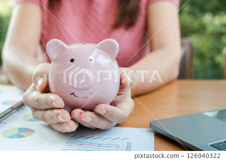 Close-up of hands holding a pink piggy bank, surrounded by financial documents and a laptop, symbolizing savings and financial planning. 126940322