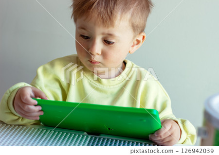 Little serious two-year-old boy in a yellow sweater playing with a green tablet at a table 126942039