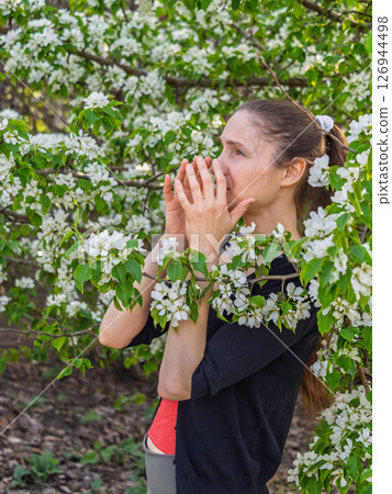 Young woman sneezes in the park against the background of a flowering tree. Allergy to pollen concept. Young woman sneezes in the park against the background of a flowering tree. Allergy to pollen concept. 126944498