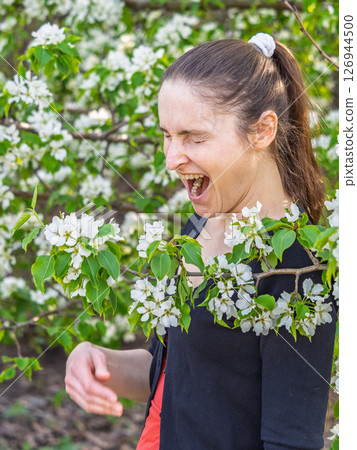 Young woman sneezes in the park against the background of a flowering tree. Allergy to pollen concept. Young woman sneezes in the park against the background of a flowering tree. Allergy to pollen concept. 126944500