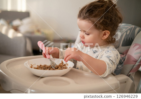 Happy toddler with curls having breakfast in a high chair. Kitchen background with bowl, spoon, and loving family environment. Happy toddler with curls having breakfast in a high chair. Kitchen background with bowl, spoon, and loving family environment. 126946229