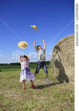Brother and sister throwing straw hats next to a big hay bale 126946689