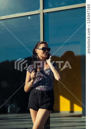 A girl wearing glasses is talking on the phone, leaning against the glass wall of a modern building 126947168