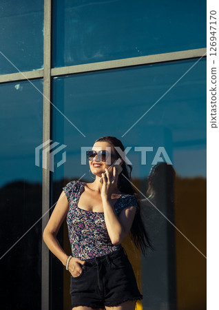 A young woman wearing shorts and a T-shirt stands near the transparent facade, holding a cell phone 126947170