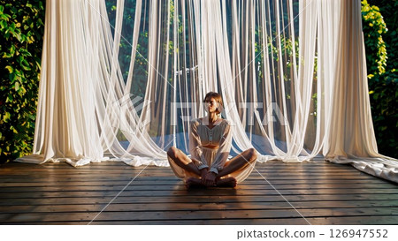 Serene yogi meditating in white curtained gazebo, surrounded by lush greenery, finding inner calm through mindful breathing and peaceful pose 126947552
