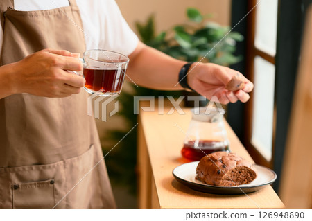 Close up of man in an apron holding a glass cup of black coffee and a piece of bread Close up of man in an apron holding a glass cup of black coffee and a piece of bread 126948890
