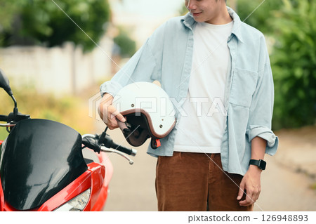 Young man standing next to a red scooter while holding helmet in one hand. Travel safety and lifestyle concept 126948893