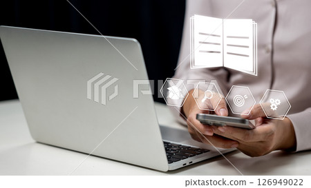 Businesswoman using a cellphone and showing a book icon. Laptop on the table. Technology innovation on a virtual screen. Concept of E-library Businesswoman using a cellphone and showing a book icon. Laptop on the table. Technology innovation on a virtual screen. Concept of E-library 126949022