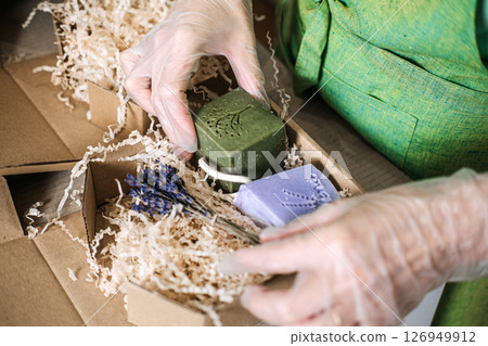 Gloved hands pack green and lavender soaps with dried lavender into a kraft gift box. Mindful shopping, slow living, ethical production, conscious consumer 126949912